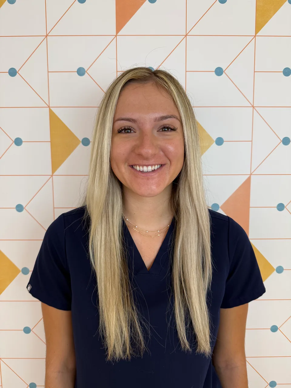 Poppy Kids registered dental assistant Olivia, smiling in her uniform in front of a patterned wall.